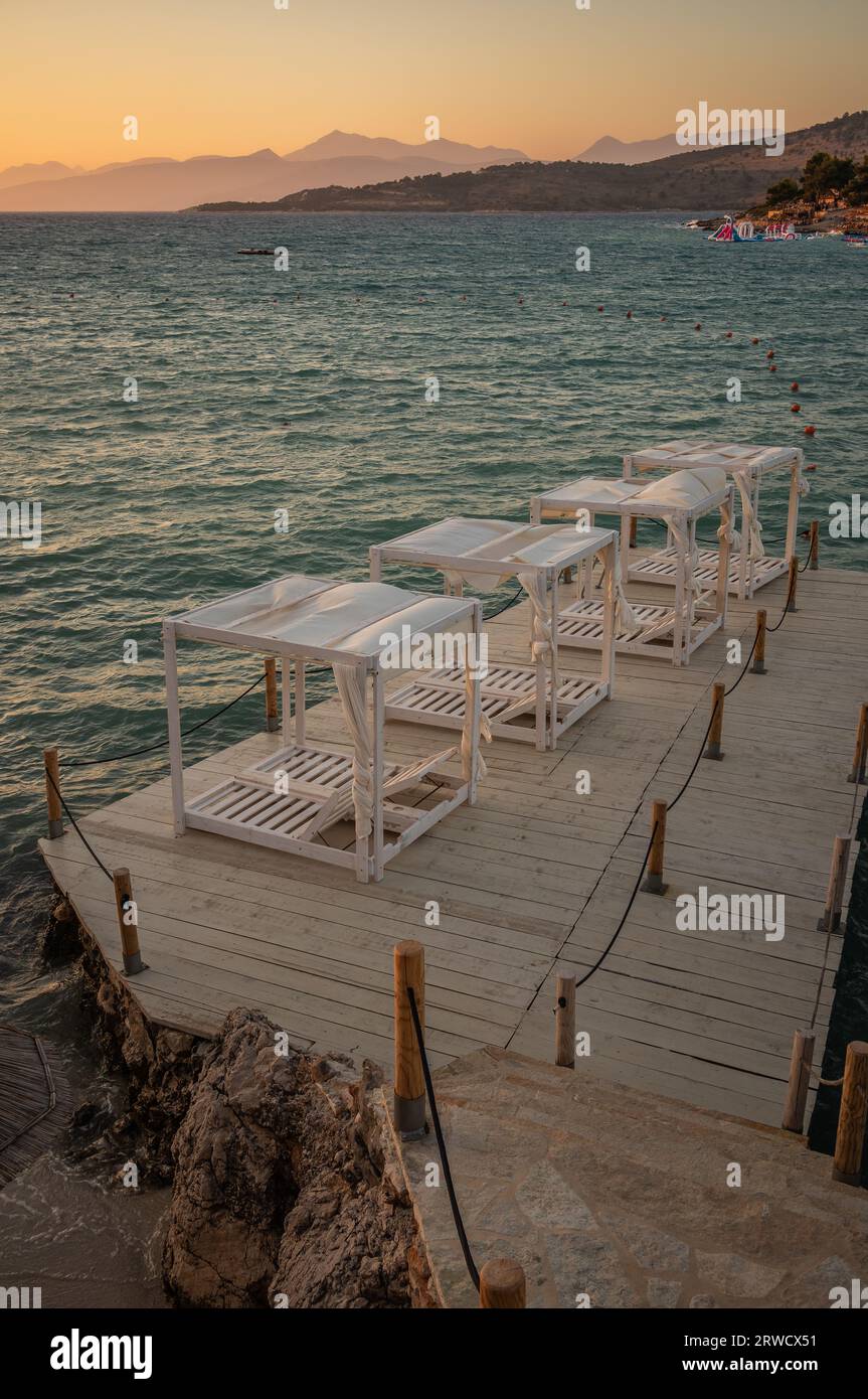 Vertikaler Blick auf den hölzernen Pier mit Liege während der Goldenen Stunde in Ksamil. Wunderschöne albanische Riviera mit Sonnenuntergangsfarben. Ionisches Meer und Berge. Stockfoto