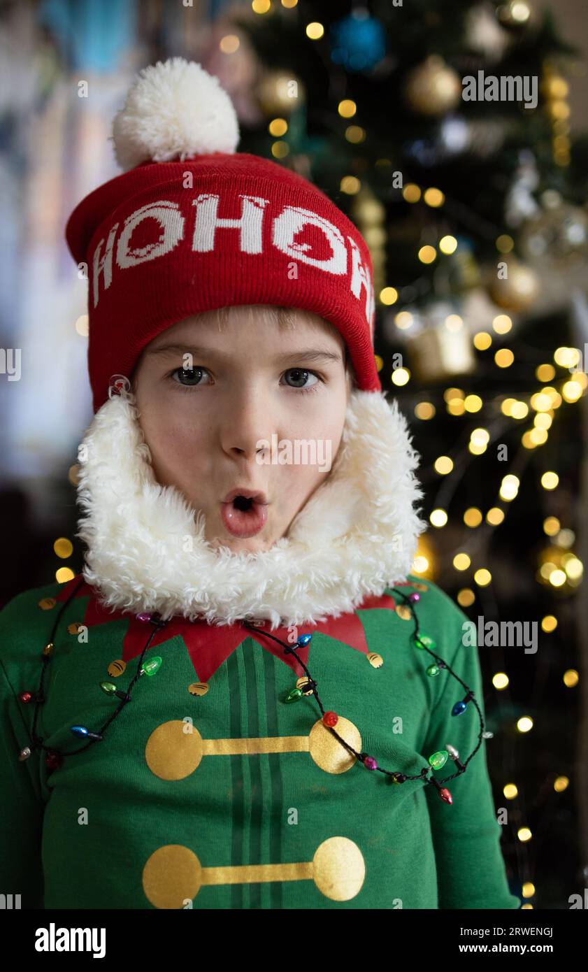 Ein Kind im Elfenkostüm und ein Weihnachtsmannhut macht Gesichter. Emotionen der Überraschung und Freude. Weihnachten, Neujahr, frohe Winterferien, Verwöhnung zu Hause. H Stockfoto