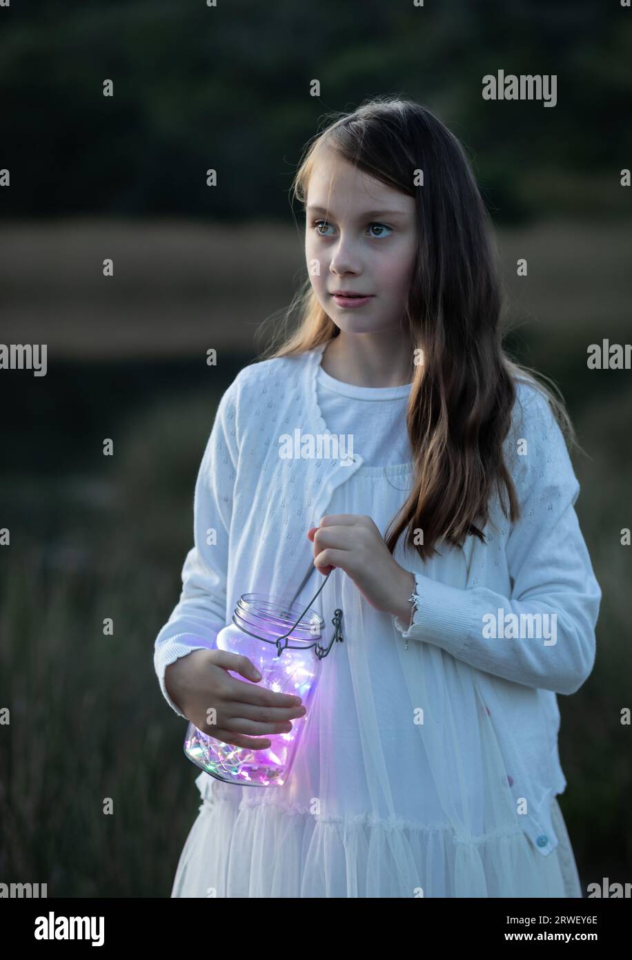 Nahaufnahme eines jungen Mädchens, das in der Abenddämmerung ein Glasgefäß mit Feenlichtern hält und zur Seite schaut Stockfoto