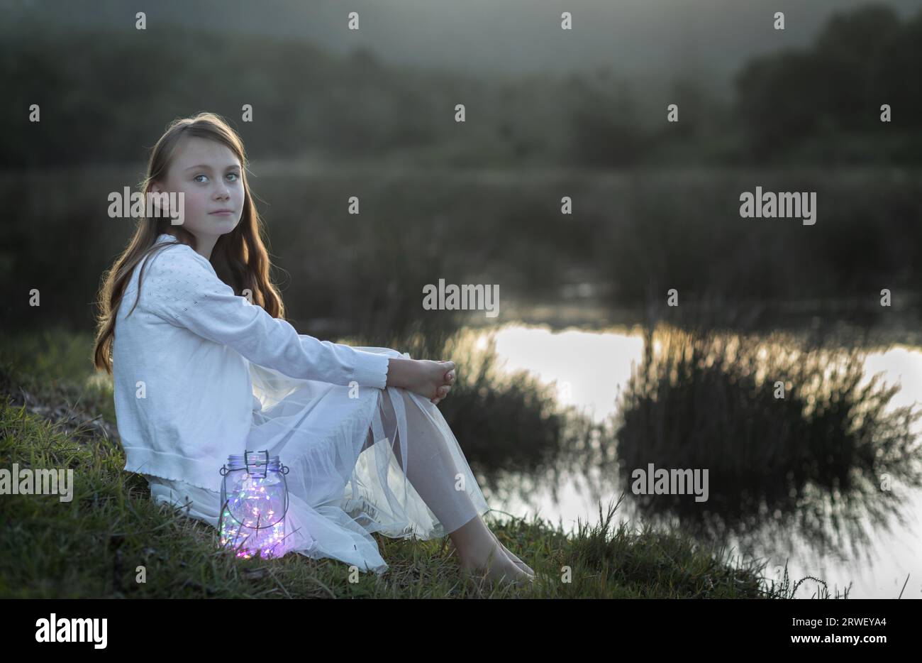 Hintergrundbeleuchtetes Porträt eines weißen Mädchens, das am Wasserrand sitzt und auf die Kamera schaut Stockfoto