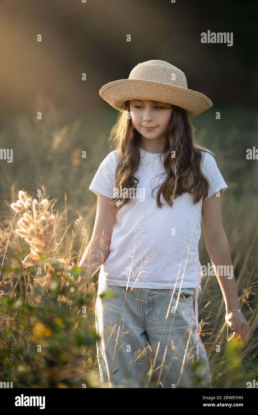 Das junge Mädchen, lässig gekleidet und mit einem Hut, steht auf einem Feld und hält ein Stück getrocknetes Gras Stockfoto