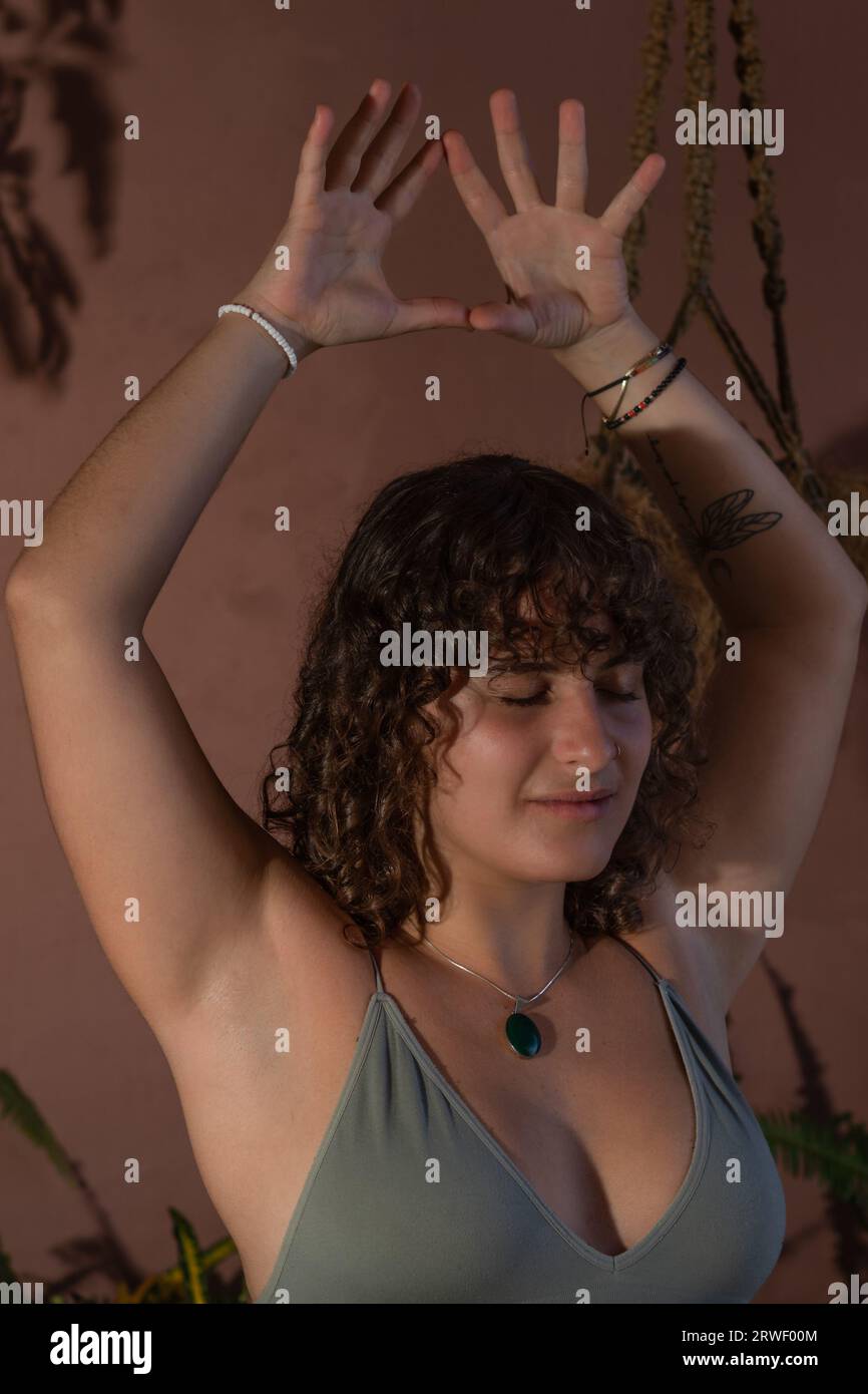 Nahaufnahme einer jungen Frau mit lockigem, dunklem Haar, die bei schwachem Licht meditiert, während ihre Arme über dem Kopf stehen Stockfoto