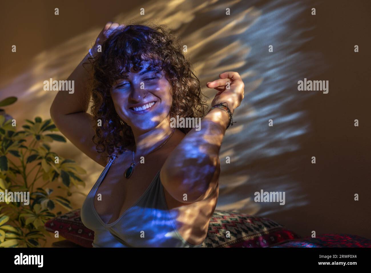 Porträt einer lächelnden jungen Frau mit lockigen dunklen Haaren, die ihre Arme in die Luft heben, als ob sie in gebrochenem Licht tanzt Stockfoto