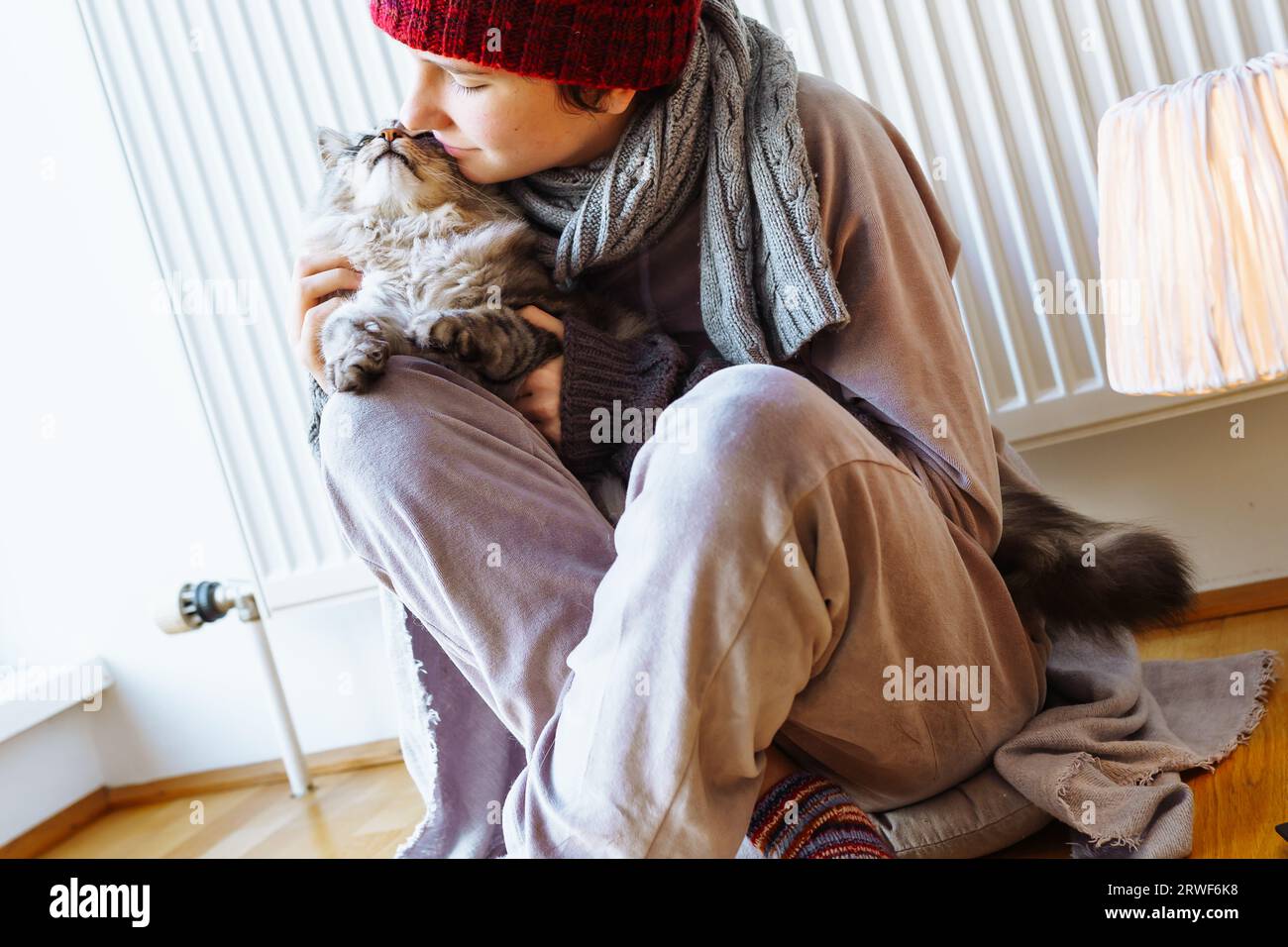 Frau mit Strickmütze, Schal, Fäustlingen, umarmter Katze, sitzt zu Hause in der Nähe des Kühlers Stockfoto