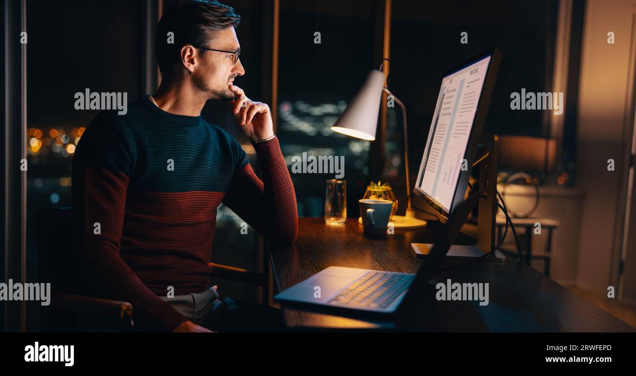 Ein kaukasischer Geschäftsmann arbeitet spät in seinem Büro und konzentriert sich auf ein Marketingprojekt. Er sitzt mit einem Laptop an seinem Schreibtisch und plant und strategisiert klug Stockfoto