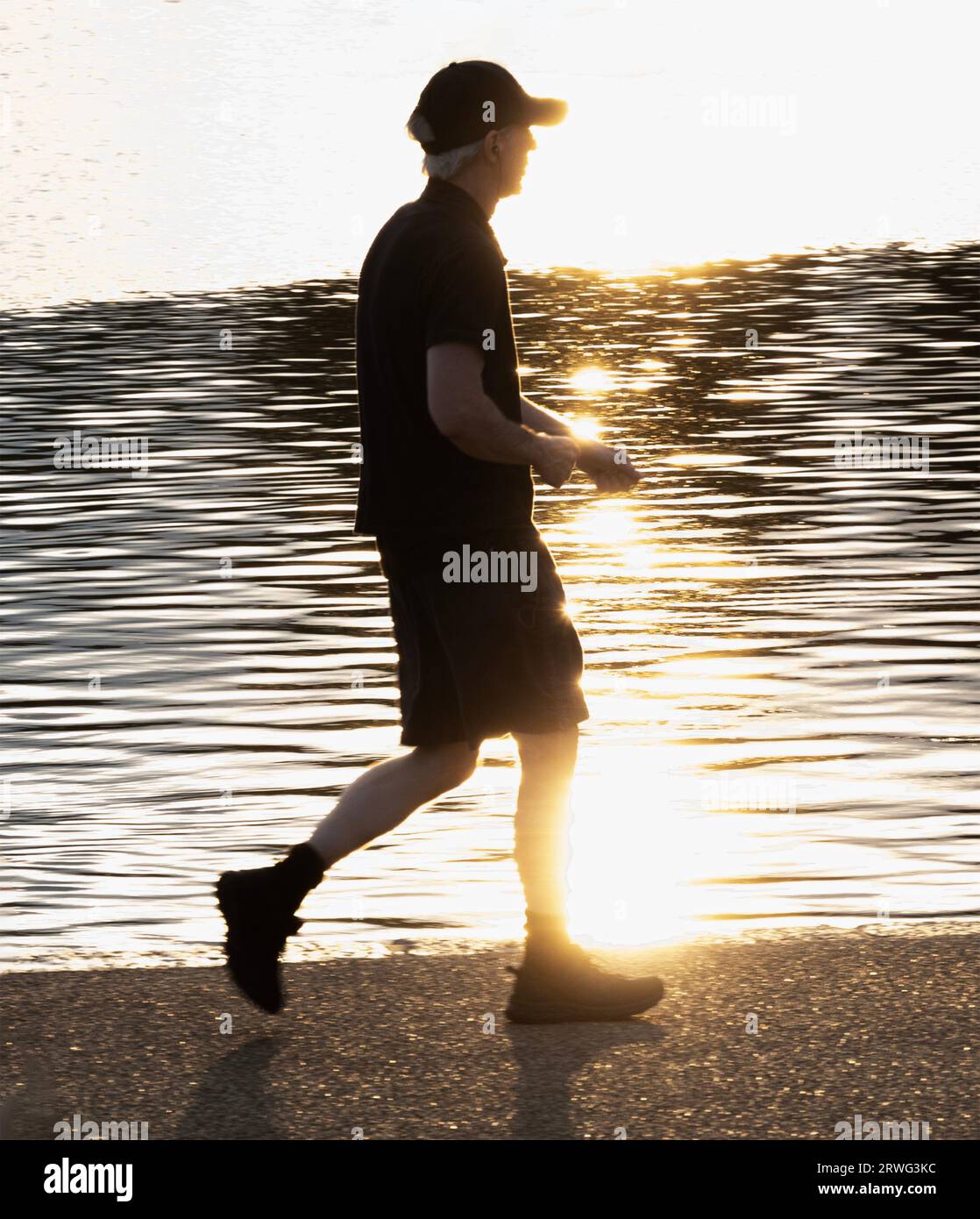 Teilweise Silhouette eines jungen Mannes in Sportausrüstung, der um den Round Pond in Kensington Gardens läuft; die Sonne ist hinter ihm, er ist Contra Jour Stockfoto