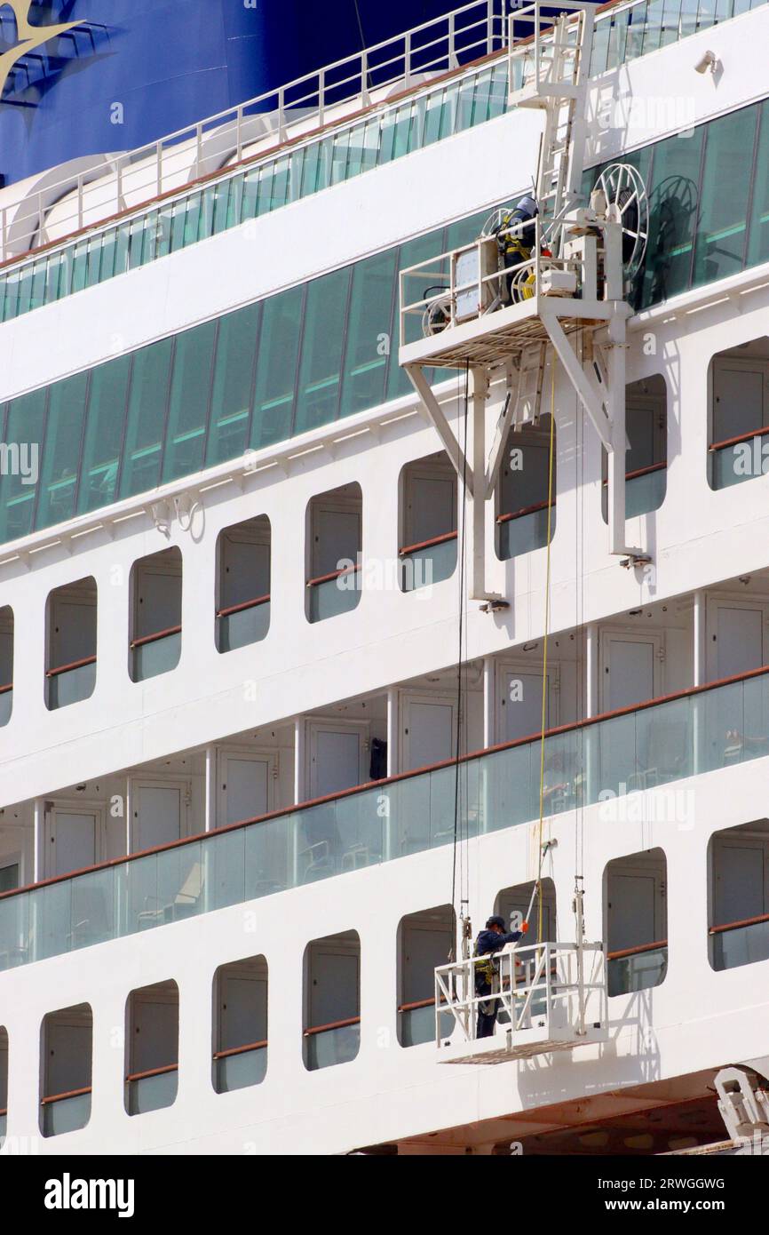 Eine Wartungsplattform senkt eine Wiege ab, damit ein Wartungstechniker den Überbau eines in Gibraltar verankerten Kreuzfahrtschiffs lackieren kann. Stockfoto