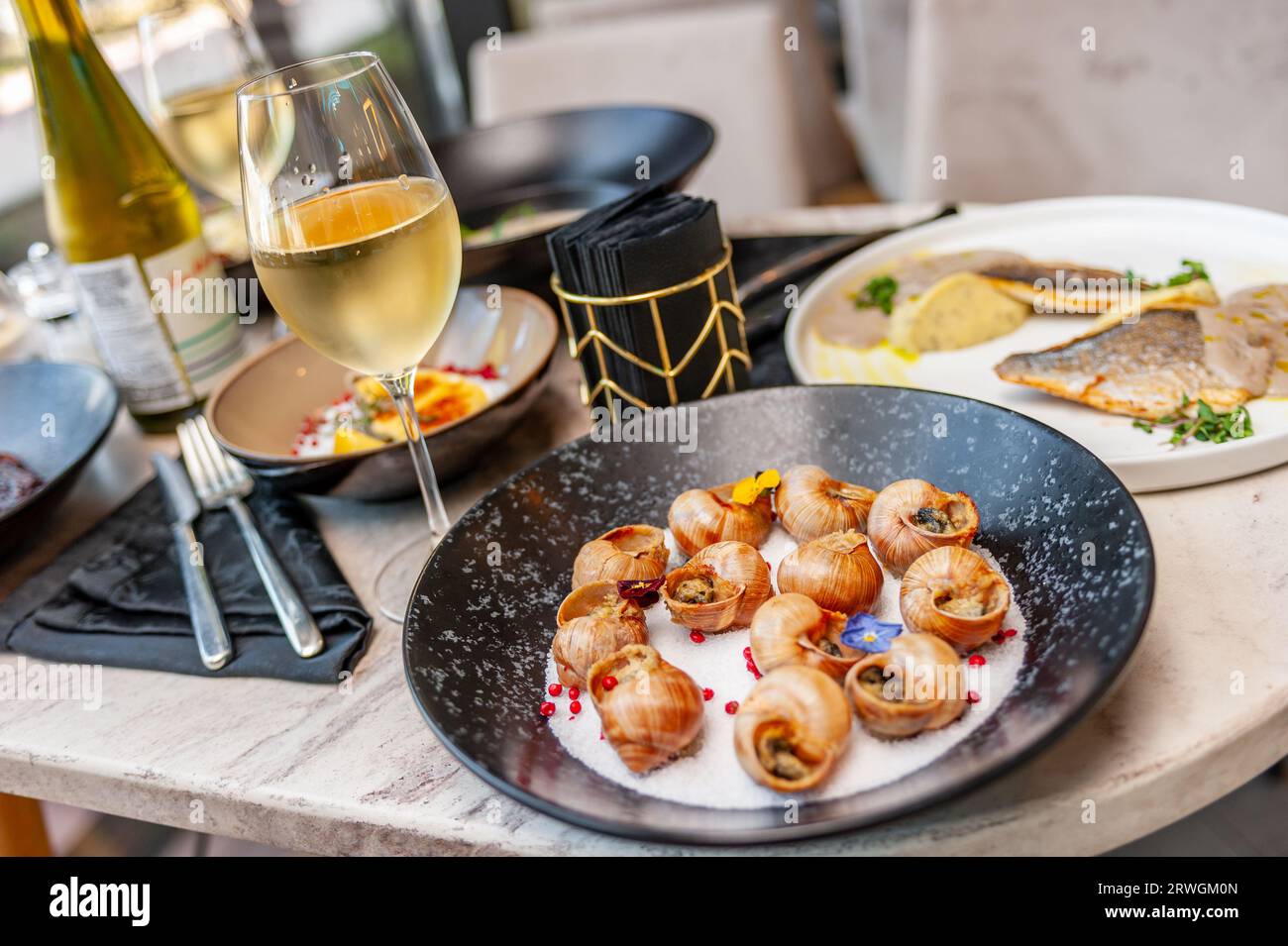 Gekochte Traubenschnecken in Muscheln, mit grünem Öl und Knoblauch auf der Oberseite gefettet, Seitenansicht auf einem Tisch in einem Restaurant mit einem Glas Weißwein Stockfoto