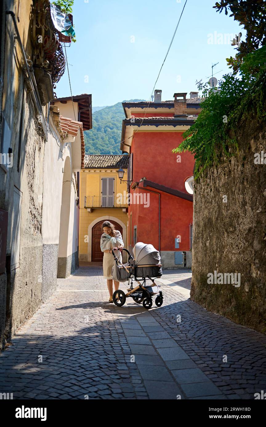 Portrait einer hübschen Frau in lässigem Kleid, die mit ihrem Baby im Kinderwagen in einer mittelalterlichen Kopfsteinpflasterstraße steht. Canzo, Lombardei, Italien Stockfoto