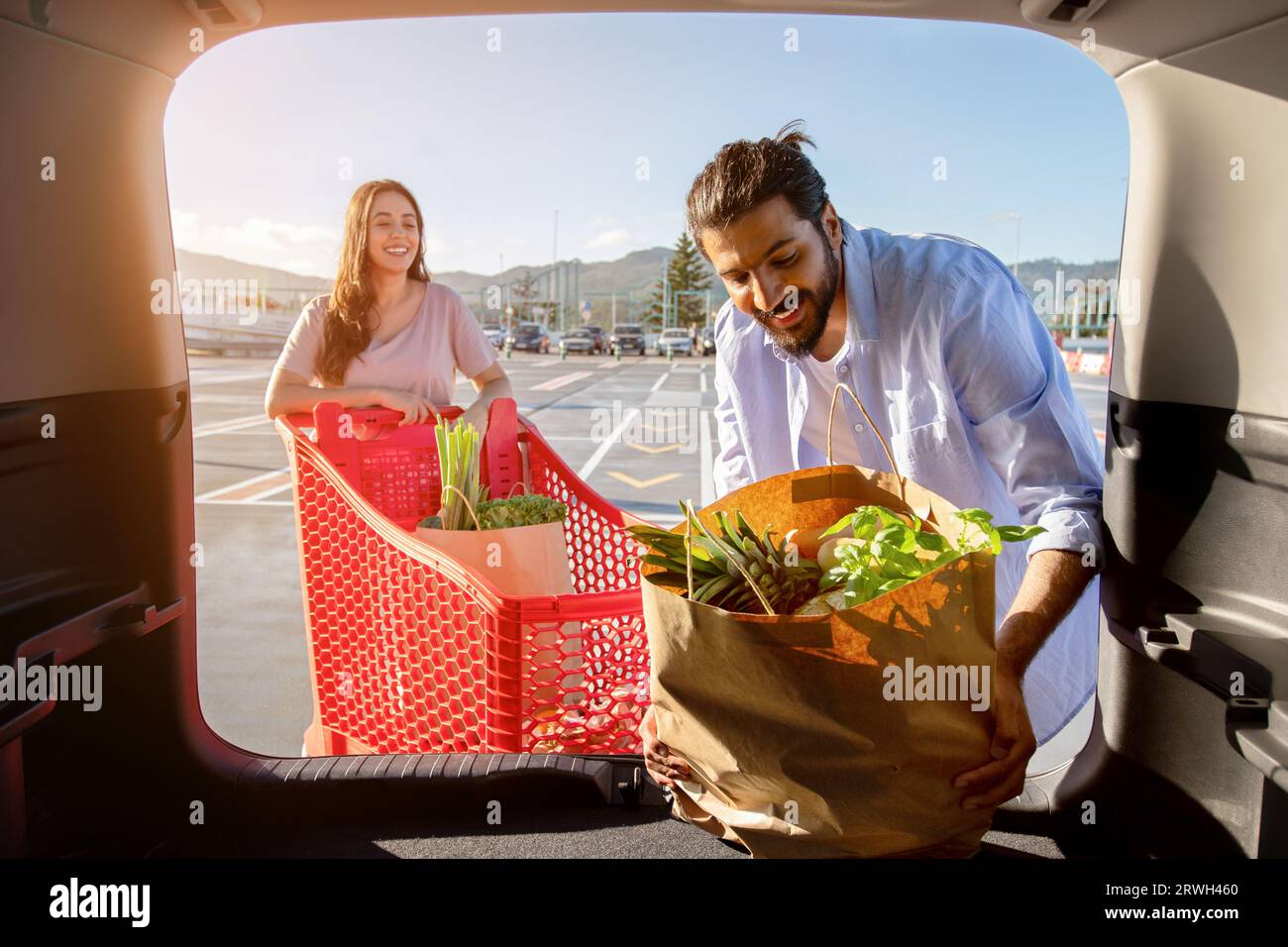 Glückliches junges indisches Paar, das Einkaufstaschen mit frischem Essen in den Kofferraum packt, Blick vom Fahrzeuginnenraum Stockfoto