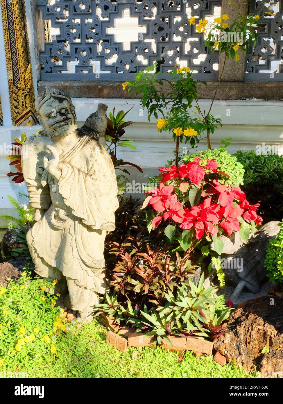 Die verwitterte Steinskulptur eines bärtigen Herrn in Gewändern, mit einem Krug auf seiner Schulter, ist eingebettet in die bunte Gartenflora gegen Stockfoto