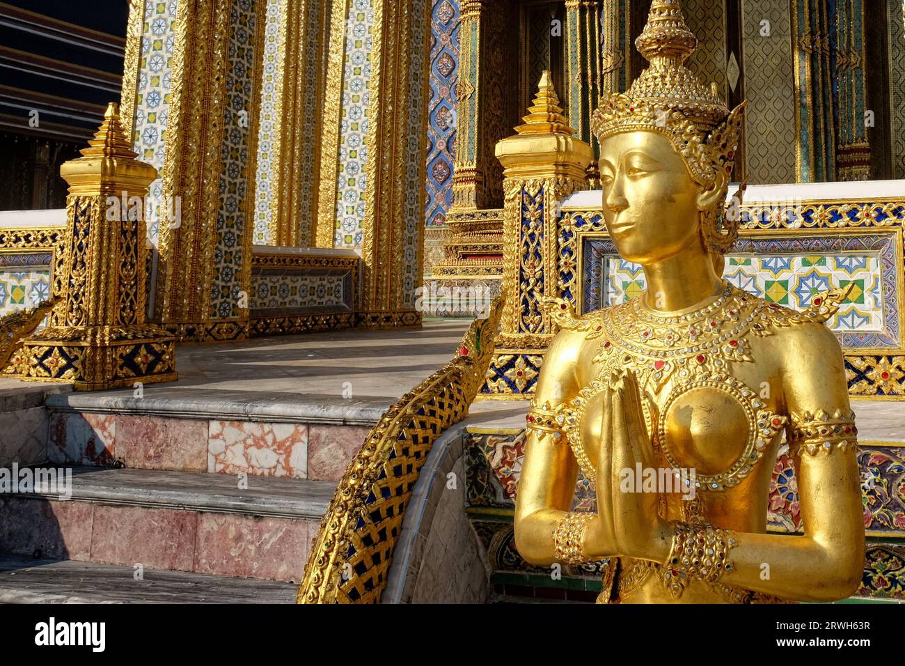 Eine goldene Statue einer Frau in traditioneller thailändischer Kleidung, vor einem Tempel mit blauen und goldenen Mosaikfliesen. Stockfoto