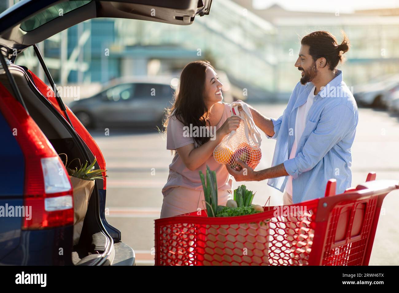 Glückliches lateinisches Paar, das auf dem Parkplatz steht und Papiertüten nach dem Einkaufen in den offenen Kofferraum lädt Stockfoto