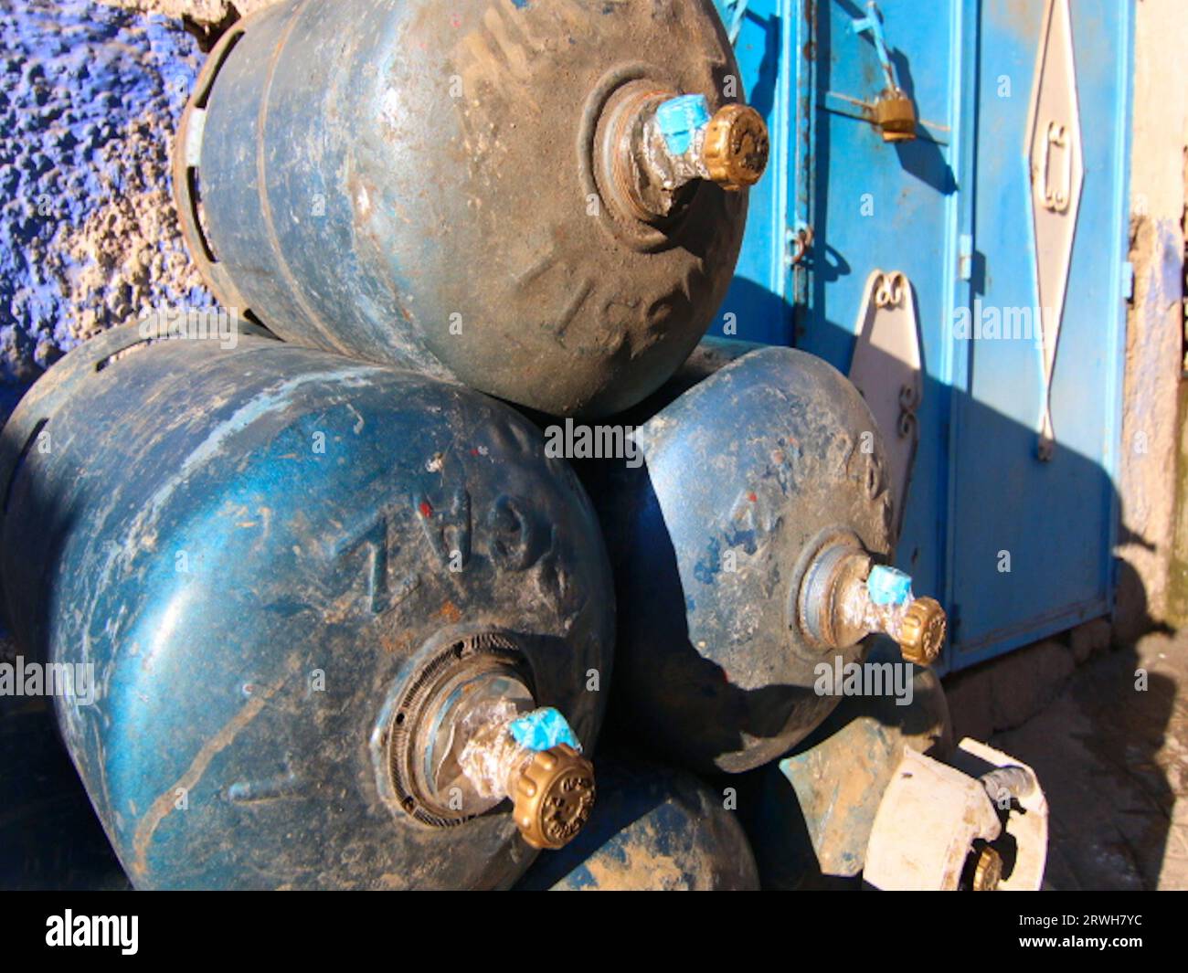 Ein Stapel von Gasflaschen aus verstörtem blauem Metall liegt an ihren Seiten übereinander, vor einer rauen strukturierten blauen Wand und gemusterten Türen. Stockfoto
