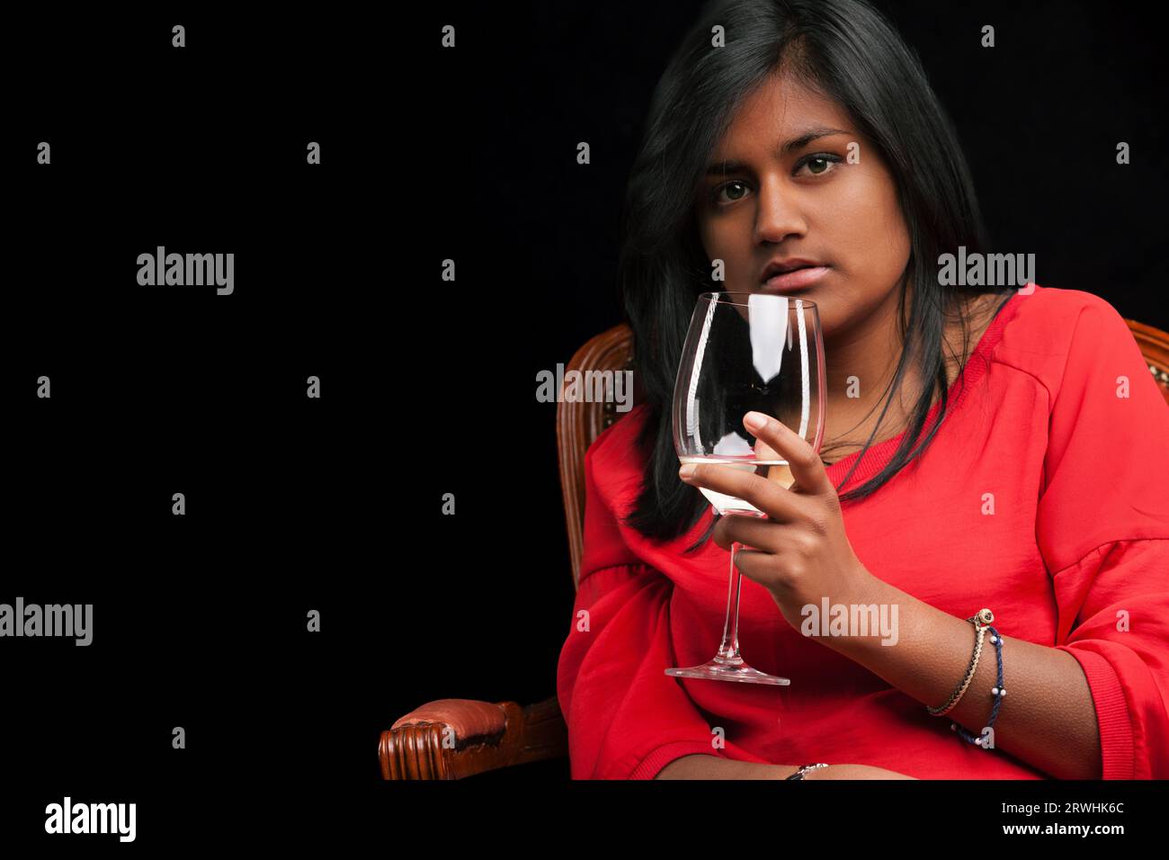 Eine indische Frau feiert die Kunst des Weins und wirbelt hochwertigen Weißwein auf einem zeitlosen Leder- und Holzstuhl. Sie ist in einem leuchtenden Rot gekleidet Stockfoto