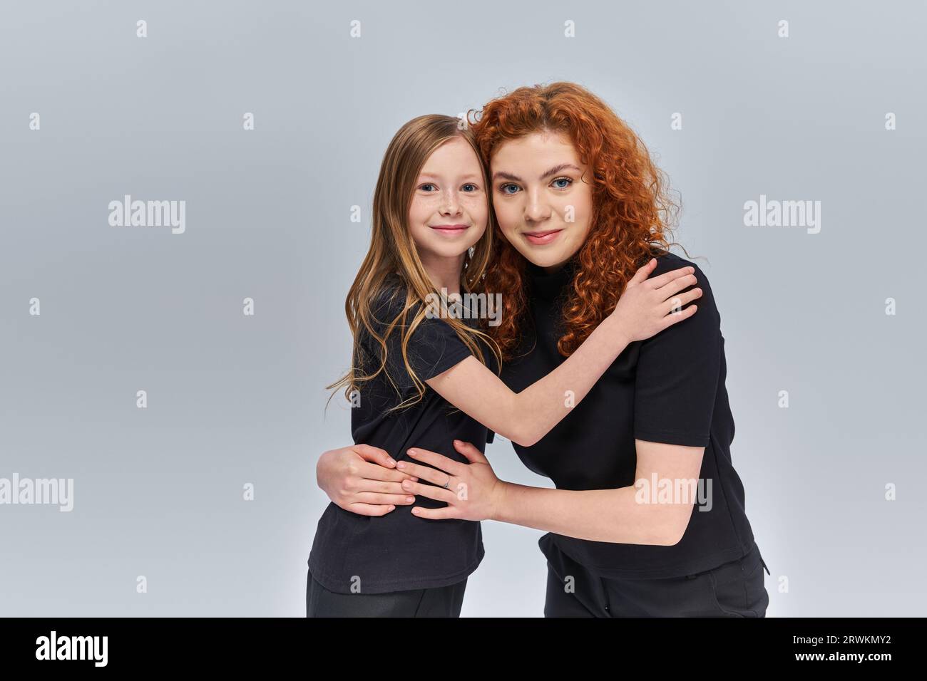 Glückliche junge Mutter mit lockigen roten Haaren, die kleines Mädchen auf grauem Hintergrund umarmen, zwei Generationen Stockfoto