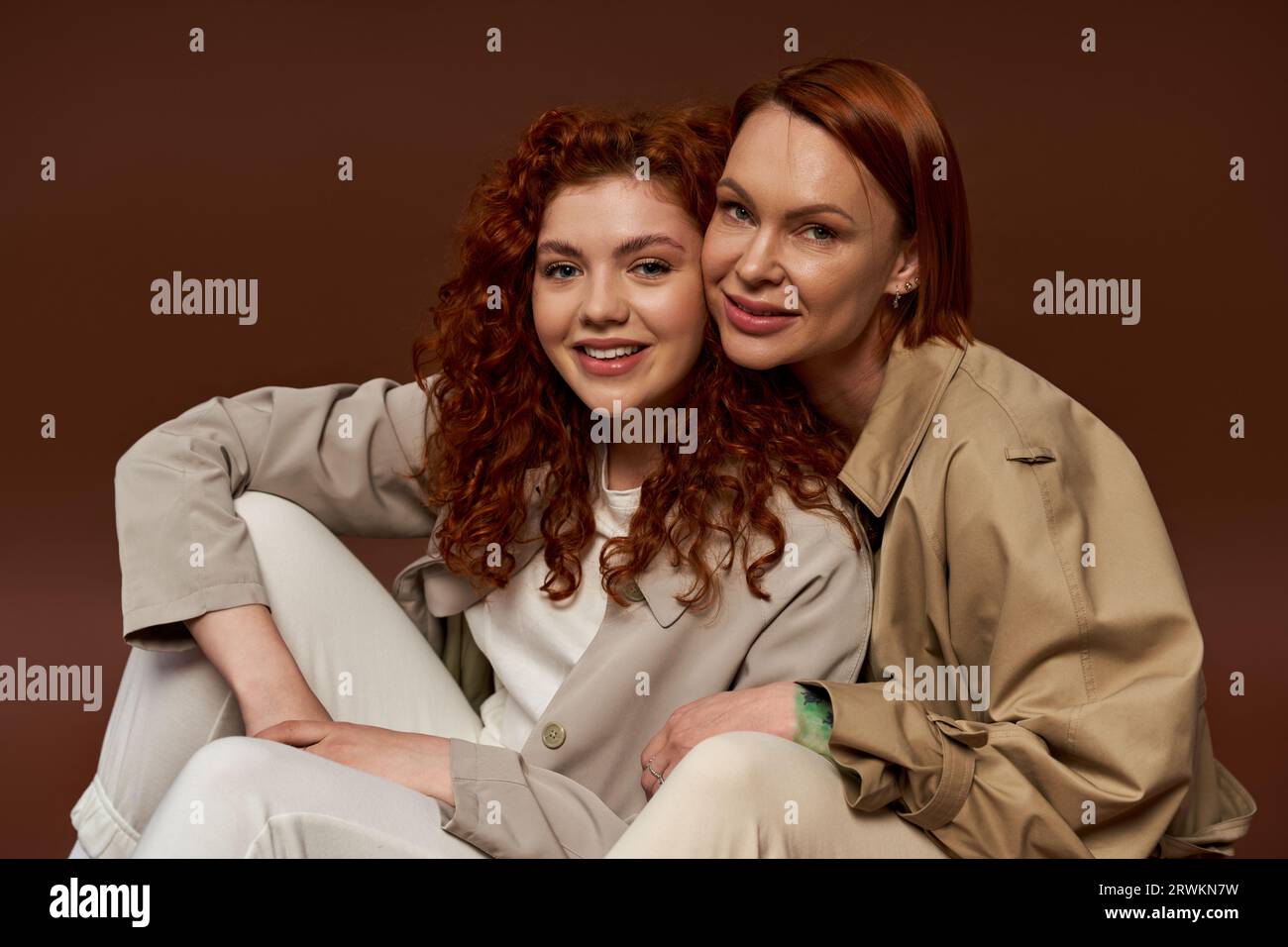 Zwei Generationen, fröhliche Frauen mit rotem Haar, die in trendiger Herbstkleidung auf braunem Hintergrund posieren Stockfoto