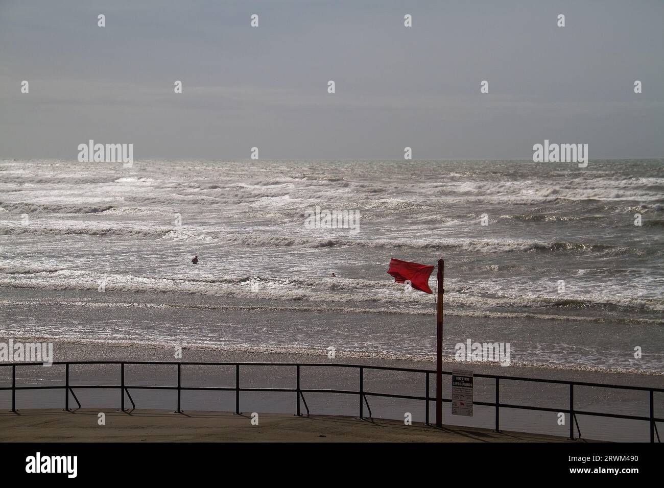 Zwei Personen in der Ferne auf einer rauen See, ignorieren eine rote Flagge Warnung vor Gefahr, dunkles Meer reflektiert letztes Sonnenlicht Stockfoto