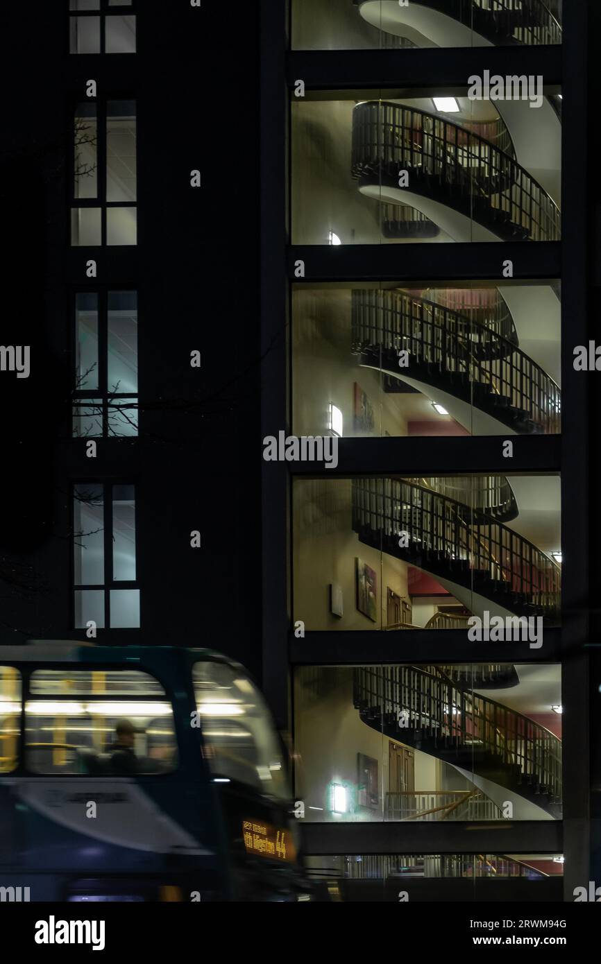 Die Hochhausarchitektur nachts zeigt eine Wendeltreppe, die durch Fenster von Etage zu Etage mit Innenbeleuchtung und Busoberseite gesehen wird Stockfoto