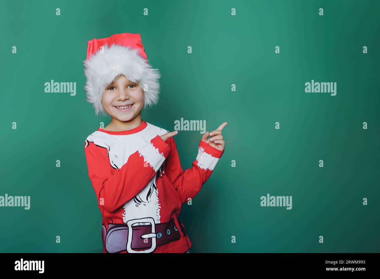 Glücklicher, lächelnder kaukasischer Junge in santa T-Shirt und Hut zeigt auf leeren Platz auf grünem Hintergrund für Werbetext. Hochwertige Fotos Stockfoto