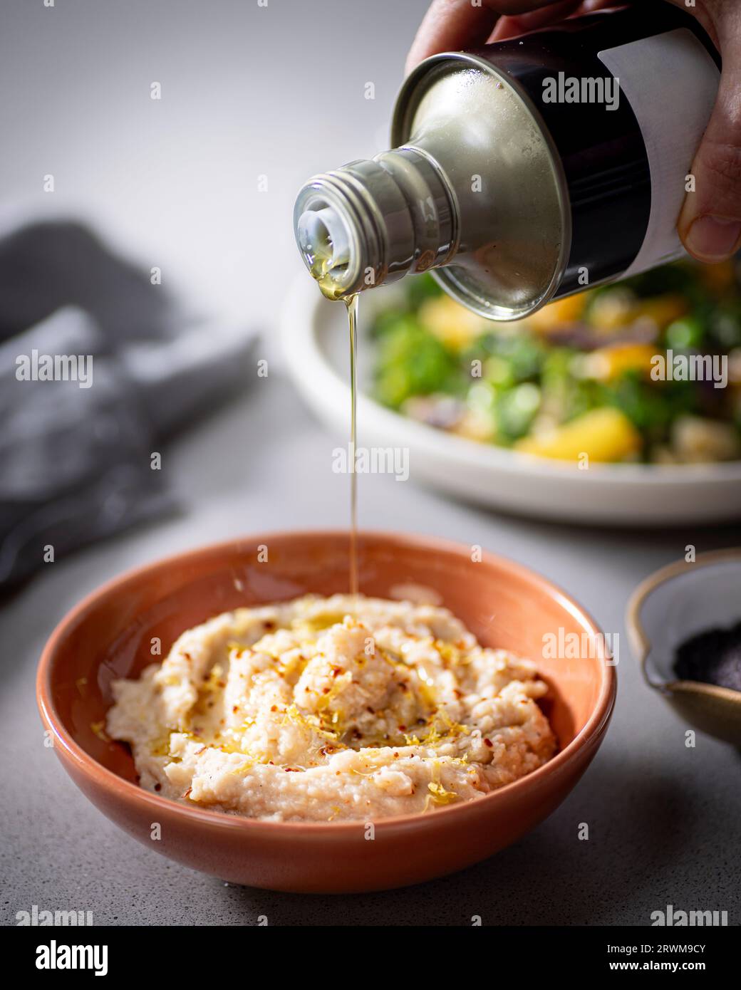 goldenes Olivenöl wird zart aus einer Flasche auf eine Schüssel gefüllt mit cremigem Hummus gegossen. Im Hintergrund ergänzt ein Teller mit frischem Gemüse den Stockfoto