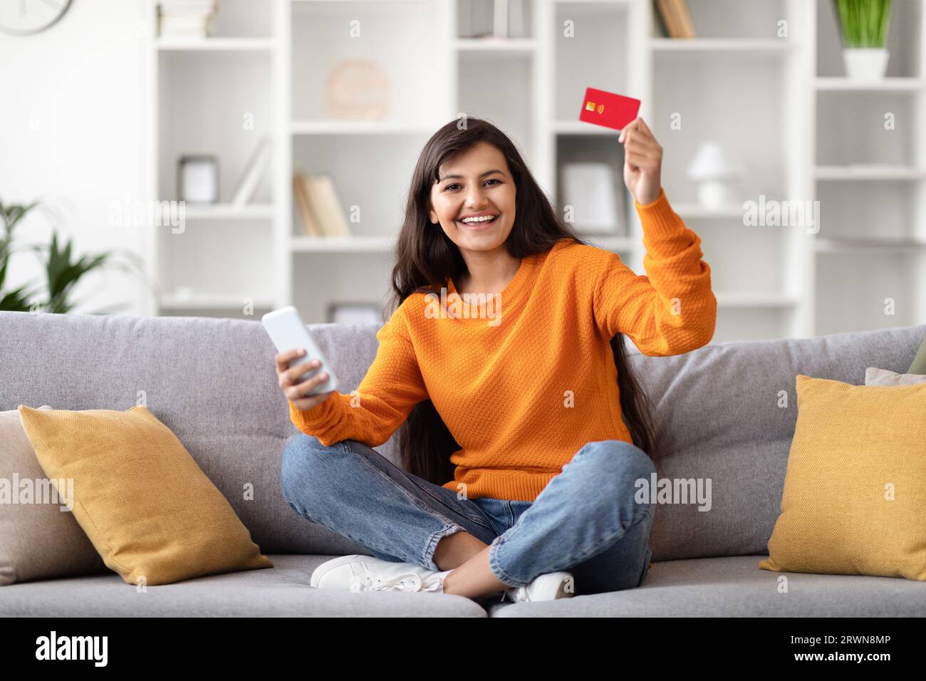 Fröhliche junge indianerin, die zu Hause Telefon und Kreditkarte benutzt Stockfoto