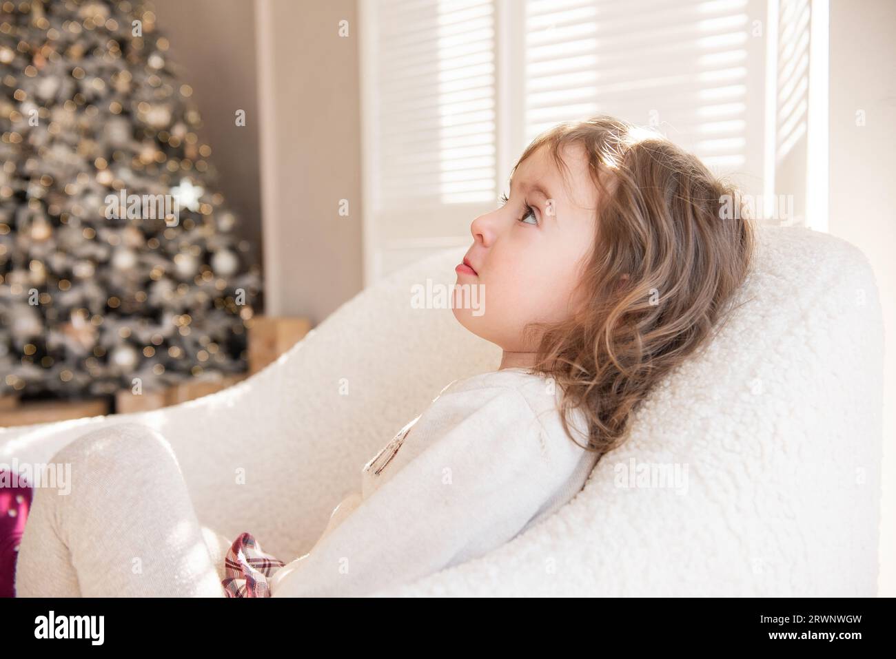 Ein nachdenkliches, träumendes kleines Mädchen liegt in einem weichen weißen Sessel und träumt von einem Geschenk des Weihnachtsmanns in der Nähe des Weihnachtsbaums. Kind in gemütlicher Atmosphäre im Stockfoto