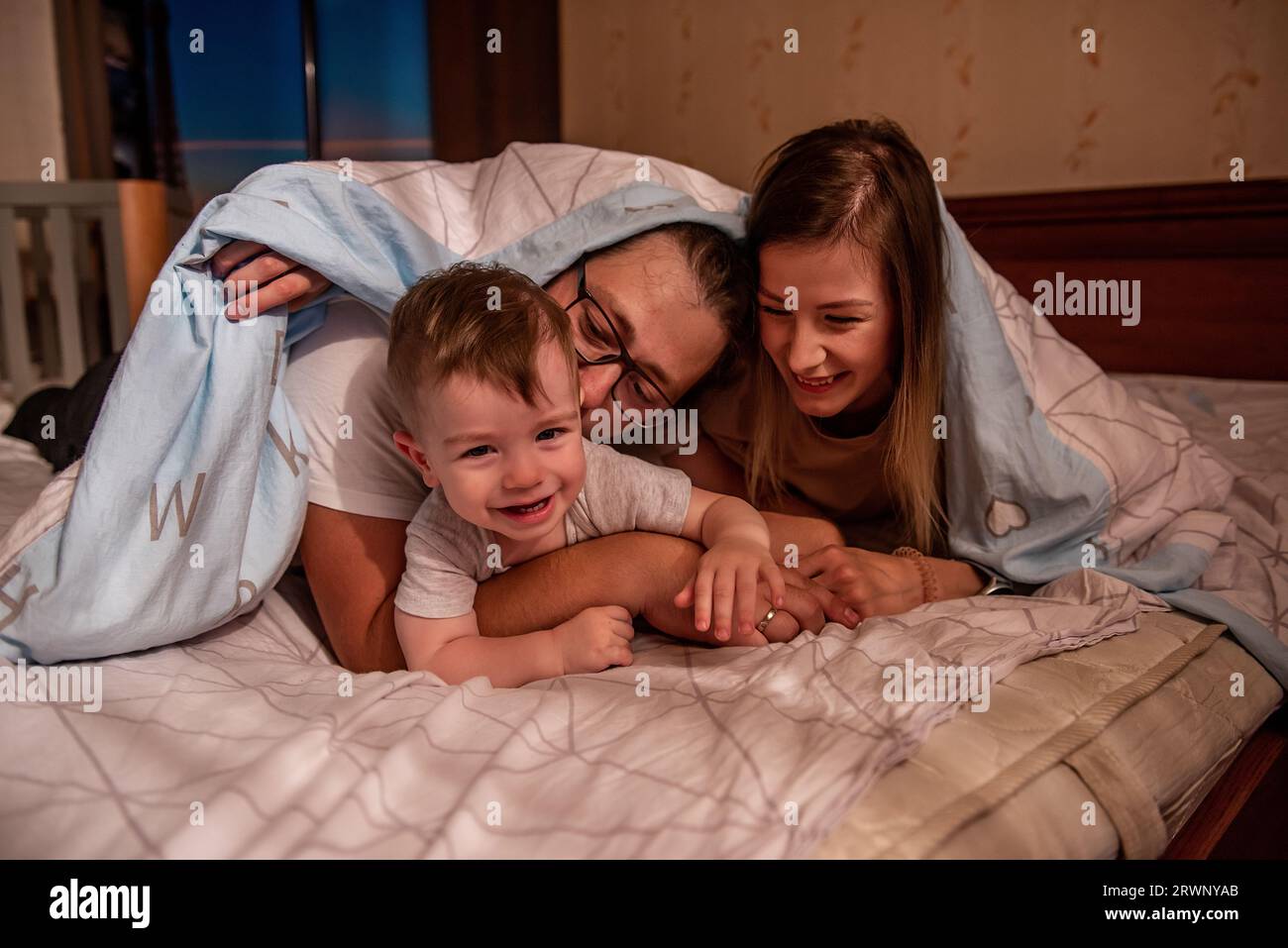 Die fröhliche, süße Familie versteckt sich mit dem kleinen Sohn unter der Decke. Mutter und Vater versuchen, ein aktives Baby zu fangen, der lustige Junge läuft vor den Eltern weg. Junge Ma Stockfoto