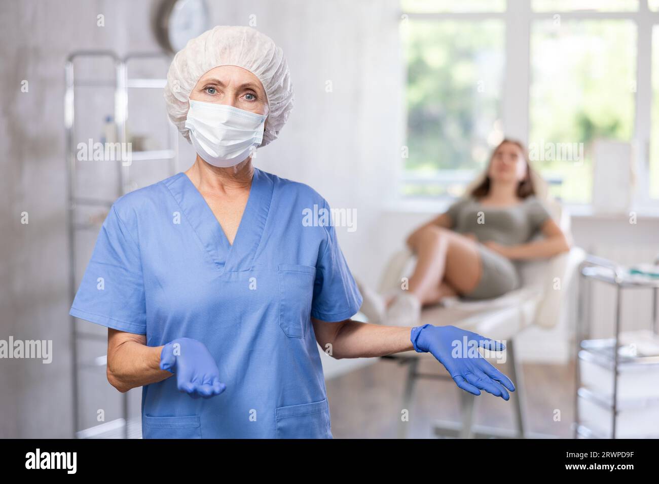 Glücklicher Arzt oder Krankenschwester mit Maske in Uniform mit gekreuztem Arm, Patient hinter dem Sofa. Stockfoto