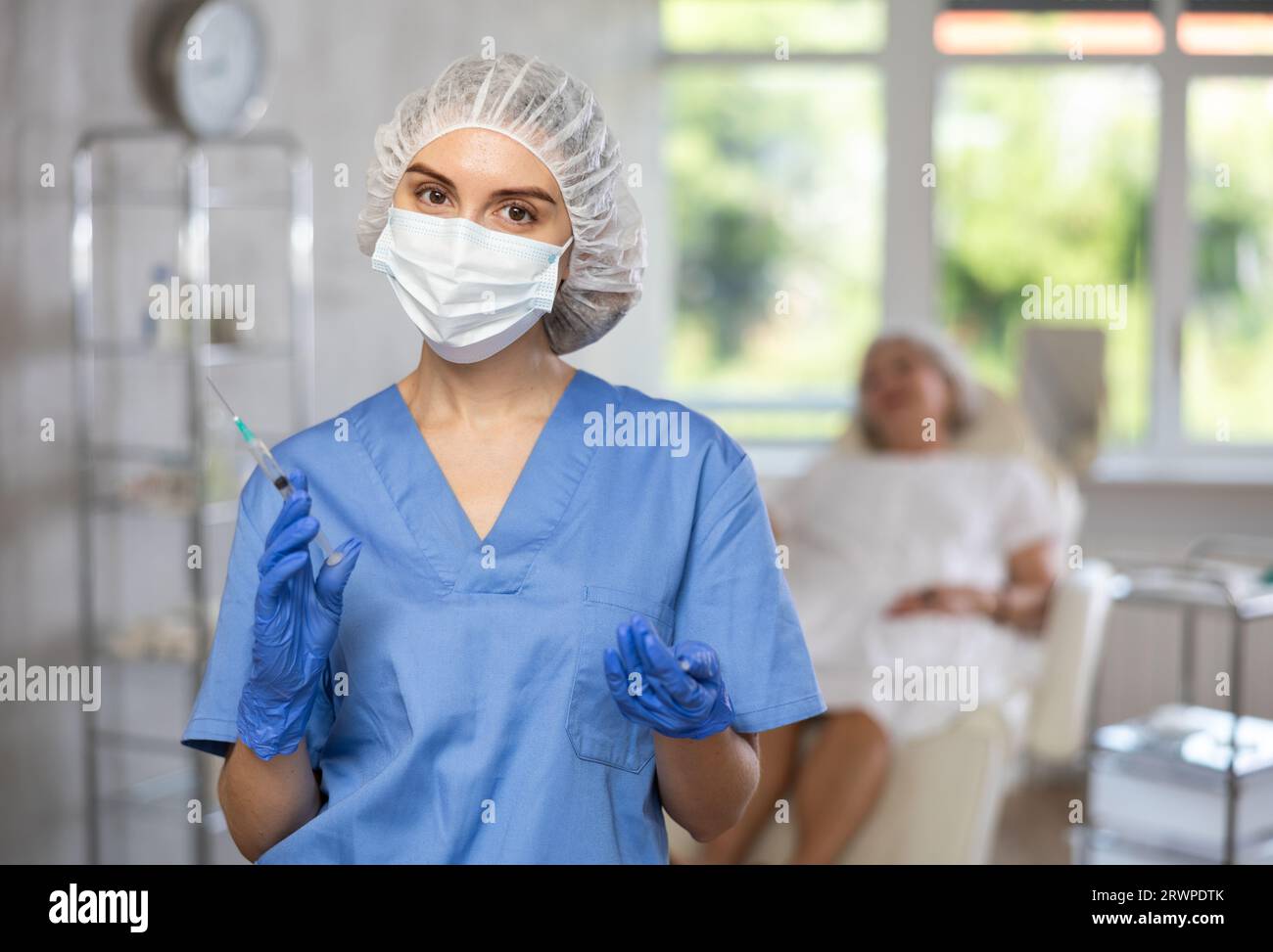 Krankenschwester in blauer Uniform mit Maske und Handschuhen hinter Hut, die Spritze mit Medizin hält, Patient dahinter. Stockfoto