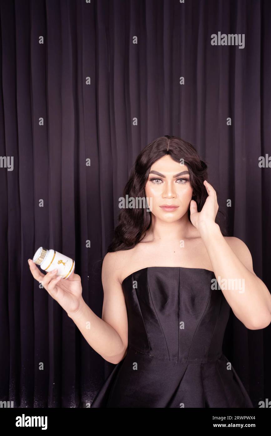 Schöne Frau, die Hautpflege in der Hand hält, während sie ein schwarzes Kleid mit schwarzen Haaren vor dem schwarzen Vorhang trägt Stockfoto