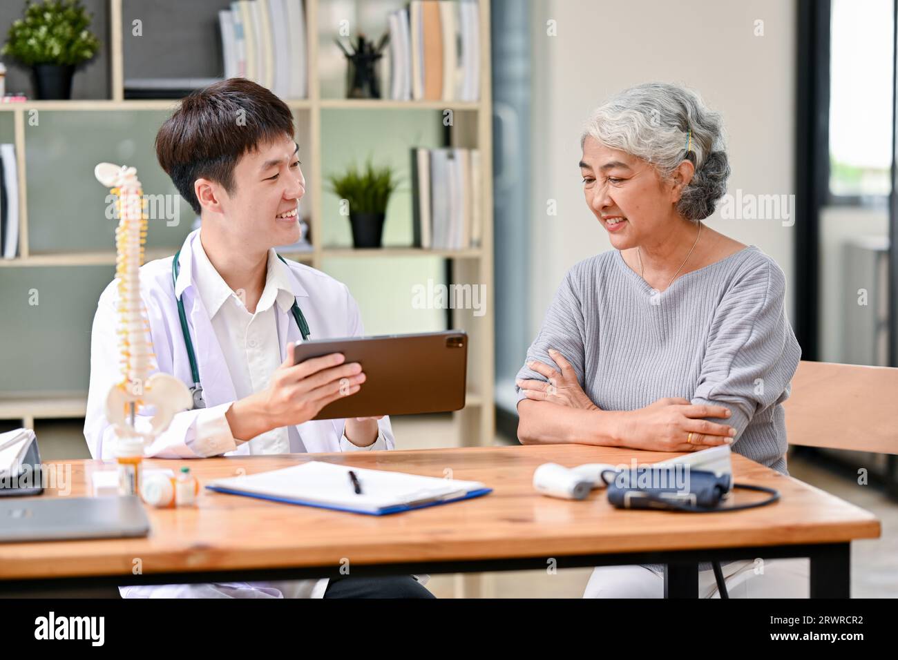 Ein fürsorglicher und freundlicher asiatischer Arzt verwendet eine digitale Tablette während einer Beratung über Behandlungspläne nach einer Operation mit einer alten Dame in einer Examina Stockfoto