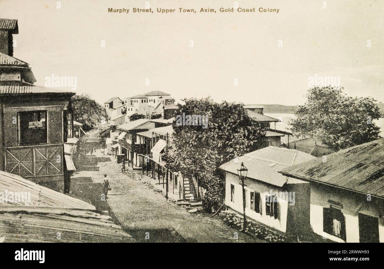 Ich kaufte eine Postkarte, die die Murphy Street in Axim, Ghana zeigt. Gebäude säumen die enge Straße und das Meer ist in der Ferne sichtbar. Gedruckte Beschriftung: Murphy Street, Upper Town, Axim, Gold Coast Colony. Stockfoto