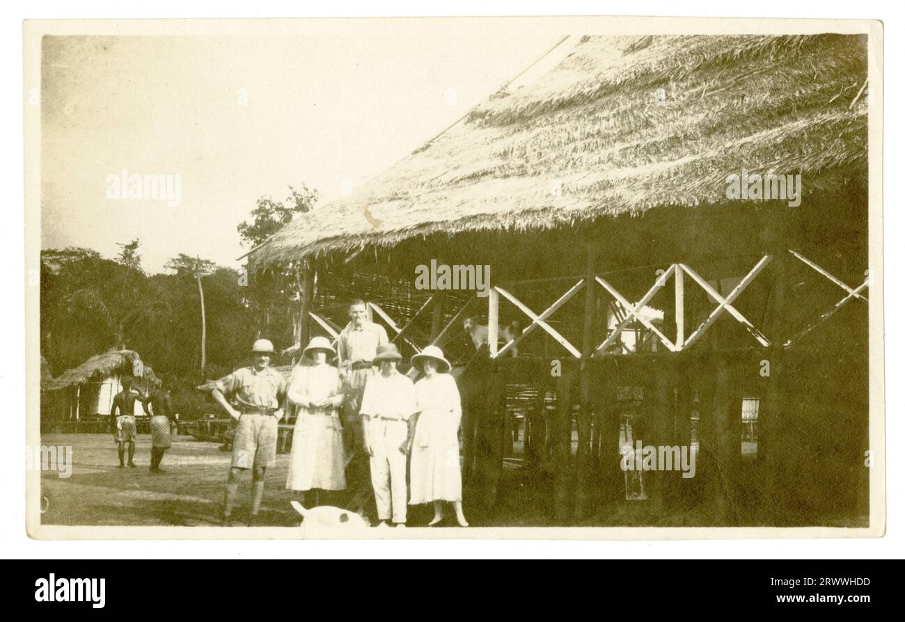 Alfred und Dorothy Tamlin stehen mit drei anderen Europäern vor einem großen Reetbau auf Stelzen mit überdachter Veranda. Es gibt ein paar afrikanische Arbeiter in der Nähe einer anderen reetgedeckten Hütte im Hintergrund. Stockfoto