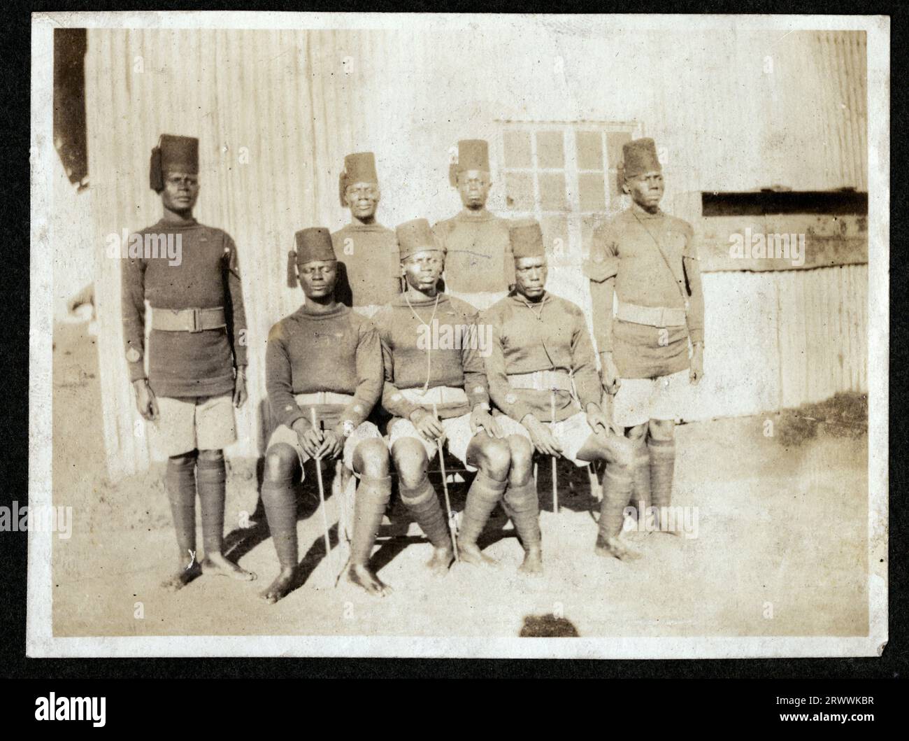 Sieben Askari aus den King's African Rifles sitzen und stehen für ein Gruppenfoto vor einem gewellten Eisengebäude. Stockfoto
