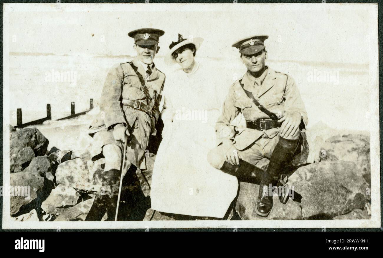 Zwei europäische Offiziere sitzen auf beiden Seiten einer elegant gekleideten Frau auf Felsen neben dem Meer in Durban, mit sichtbaren Wellen dahinter. Einer der Männer ist Bertie Rand. Originaltitel: Entlang des Strandes in Durban. Stockfoto