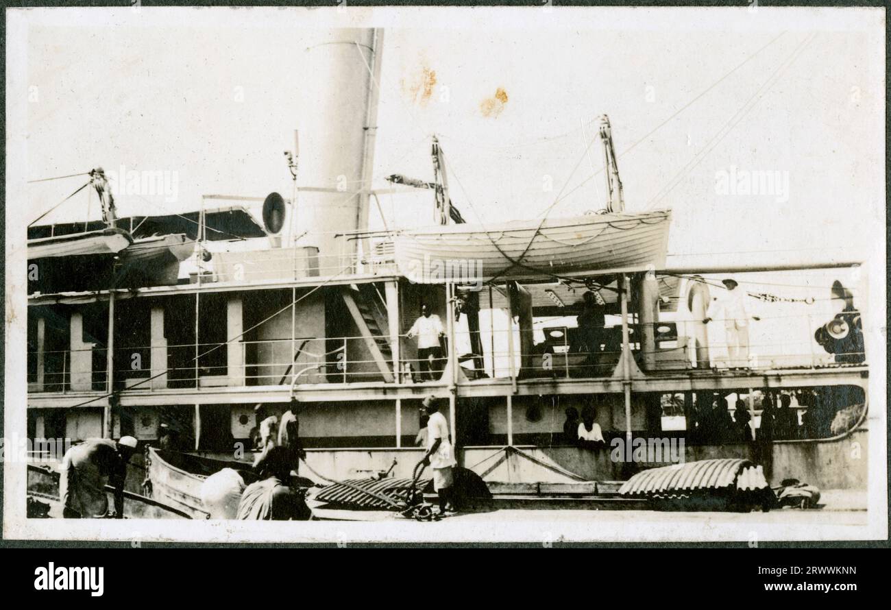 Seitenansicht des Küstendampfers SS IPU im Hafen, mit einem europäischen Offizier an Deck und afrikanischen Arbeitern, die auf Decks stehen und Fracht laden. Originaltitel: SS IPU. Stockfoto