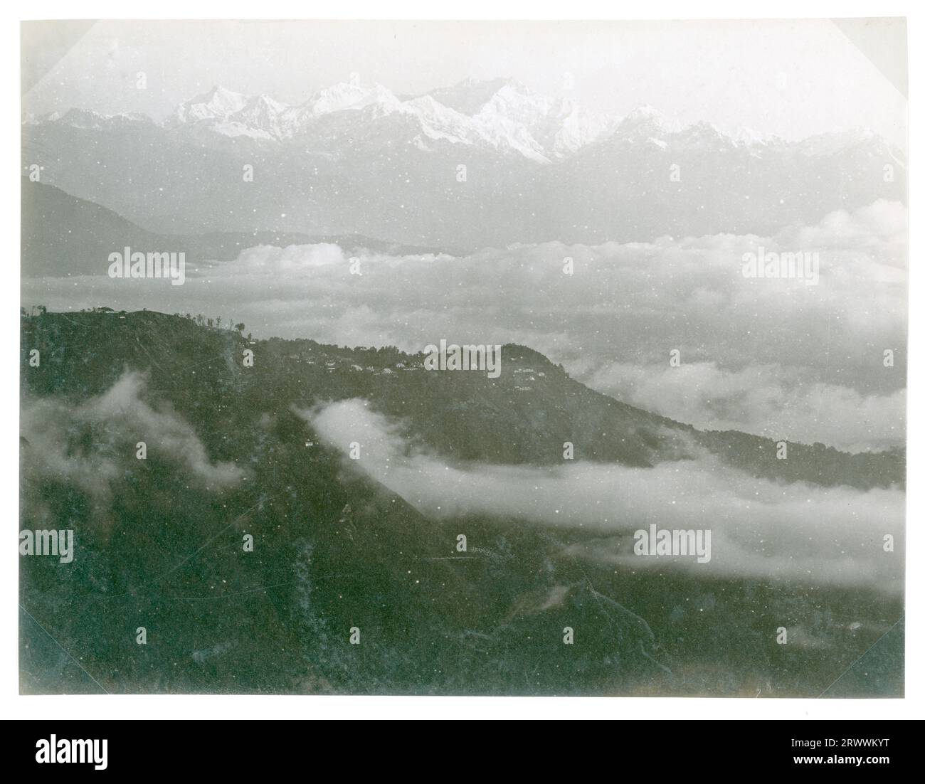 Berglandschaft mit verschneiten Gipfeln des Kangchenjunga in der Ferne. Wolken umgeben den Fuß des Berges. Die Bildunterschrift lautet: Kinchinjunga, Darjeeling im Vordergrund, vom Berg Senchal. Wolken steigen aus dem Tal auf. Stockfoto