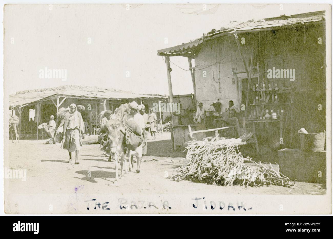 Das Foto zeigt eine Marktszene mit Ständen, Turban und anderen männlichen Figuren, eine begleitet einen schwer beladenen Esel. Die handgeschriebene Bildunterschrift bezieht sich auf die Szene als Basar, Jiddah. Stockfoto Das Foto zeigt eine Marktszene mit Ständen, Turban und anderen männlichen Figuren, eine begleitet einen schwer beladenen Esel. Die handgeschriebene Bildunterschrift bezieht sich auf die Szene als Basar, Jiddah. Stockfoto