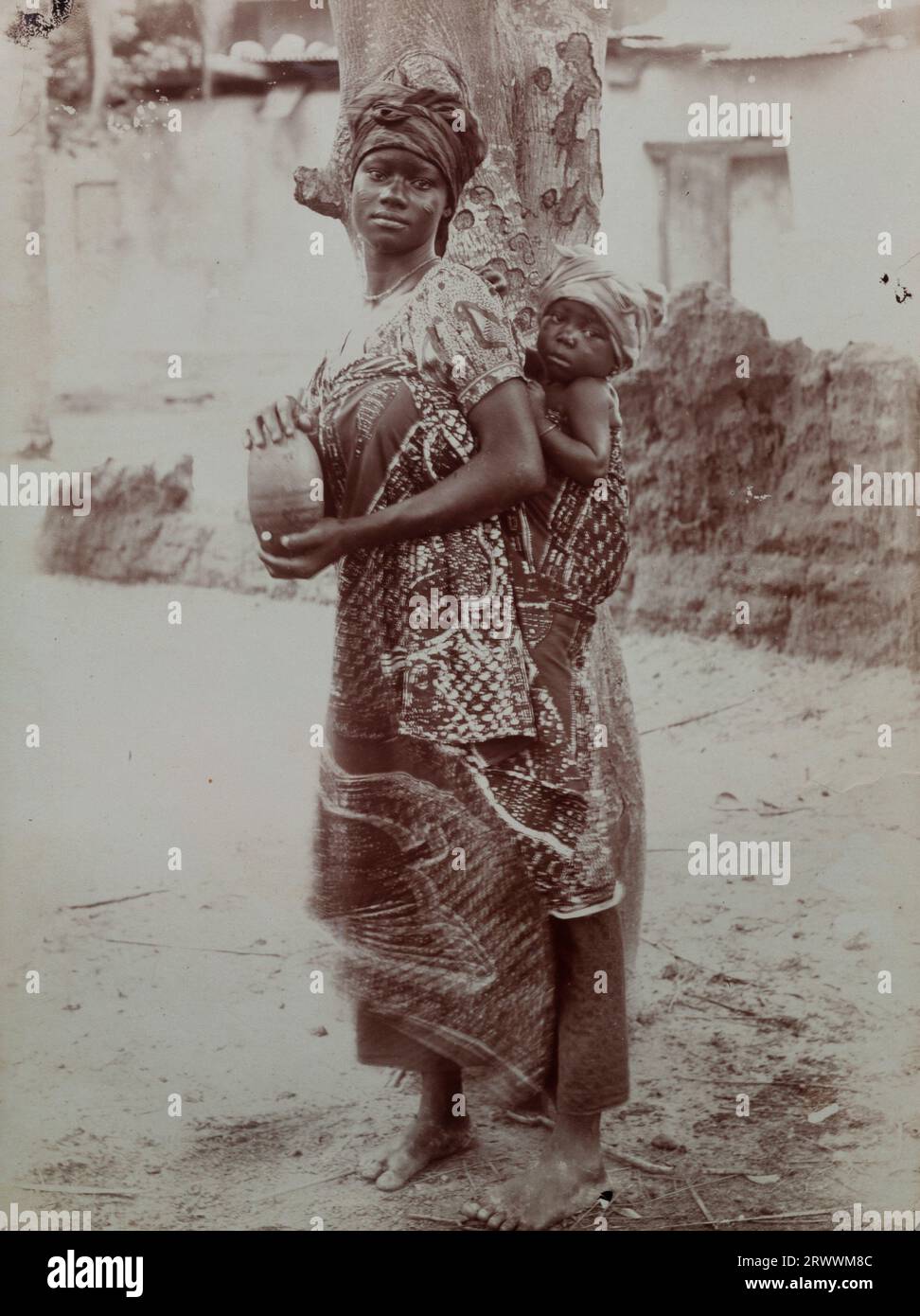 Eine Afrikanerin steht neben einem Baum und trägt ein Wickelkleid, das ein kleines Kind auf dem Rücken hält. Das Kind ist etwa ein Jahr alt. Beide tragen Kopfbedeckungen und die Frau hält einen Topf. Häuser sind hinter ihr zu sehen. Stockfoto
