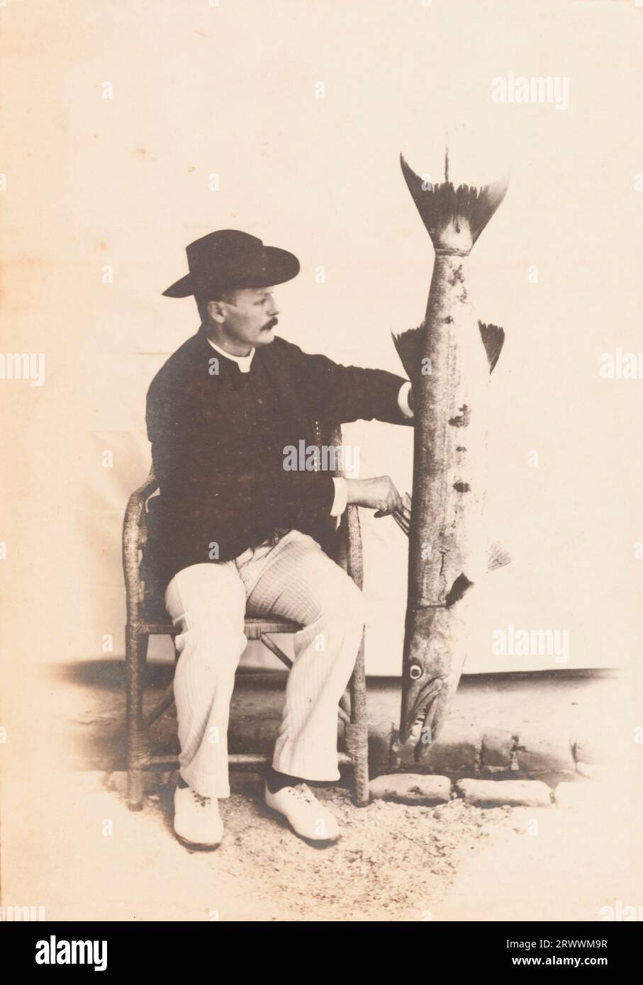 Ein europäischer Mann, der eine gestreifte Hose, eine schwarze Jacke und einen Fedora-Hut trägt, sitzt auf einem Stuhl, möglicherweise in einem Studio. Er hält einen großen Hecht auf der linken Seite auf den Kopf. Stockfoto