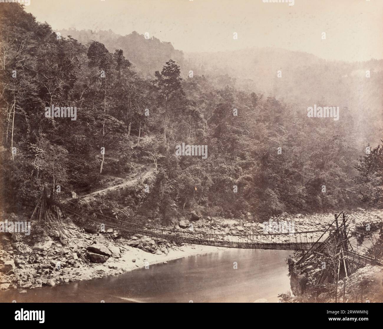 Eine Szene, die eine Bambusseilbrücke über den Rangeet River zeigt. Große Steine befinden sich auf dem Flussbett und der Hügel ist dicht mit Bäumen. Ein kleiner Pfad führt den Hügel abseits der Brücke hinauf. Die Bildunterschrift lautet: Bambusbrücke über das Rungeet. Stockfoto