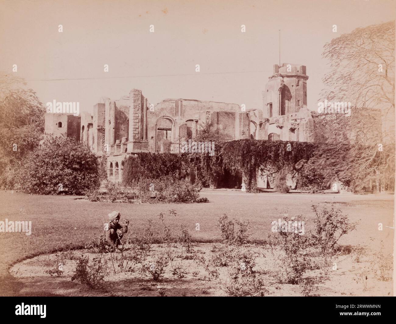Blick auf die Ruinen der britischen Residenz mit überwachsenen Pflanzen. Sie wurde während der indischen Rebellion von 1857 ruiniert. Im Vordergrund stehen Landschaftsgärten und ein junger indischer Junge hockt an einem Blumenbeet. Der Titel lautet: Lucknow Residency. Stockfoto