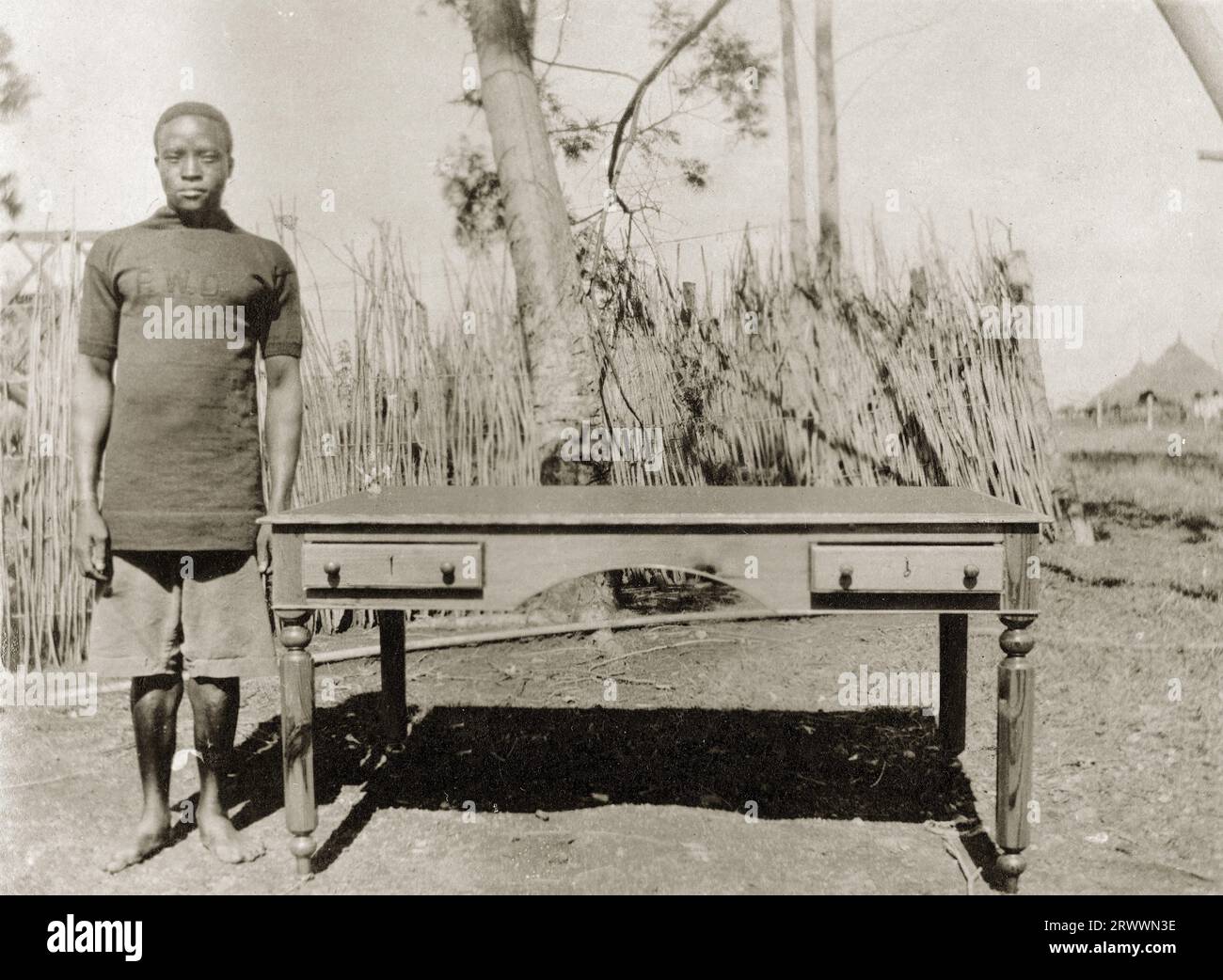 Lehrling der Abteilung für öffentliche Arbeiten, der neben einem Küchentisch mit kleinen Schubladen und gedrehten Beinen steht. Eine weitere Kopie nennt den Mann Witaba wa Sitamani und nennt das Datum März 1916. Stockfoto