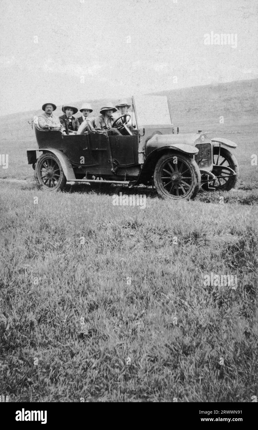 Fünf europäische Männer mit Helmen sitzen in einem offenen Tourenwagen auf einer grasbewachsenen Ebene. Einer hat ein Gewehr. Originalhandschrift: Über dem Veldt. Nakuru. Stockfoto