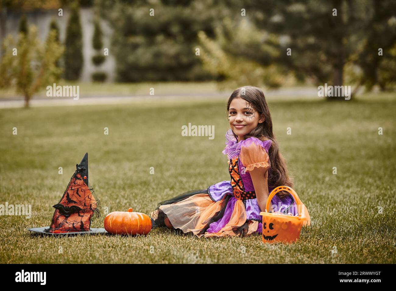 Glückliches Mädchen im Halloween Kostüm in der Nähe von dekorativem Kürbis, Spitzmütze und Süßigkeiteneimer auf Gras Stockfoto