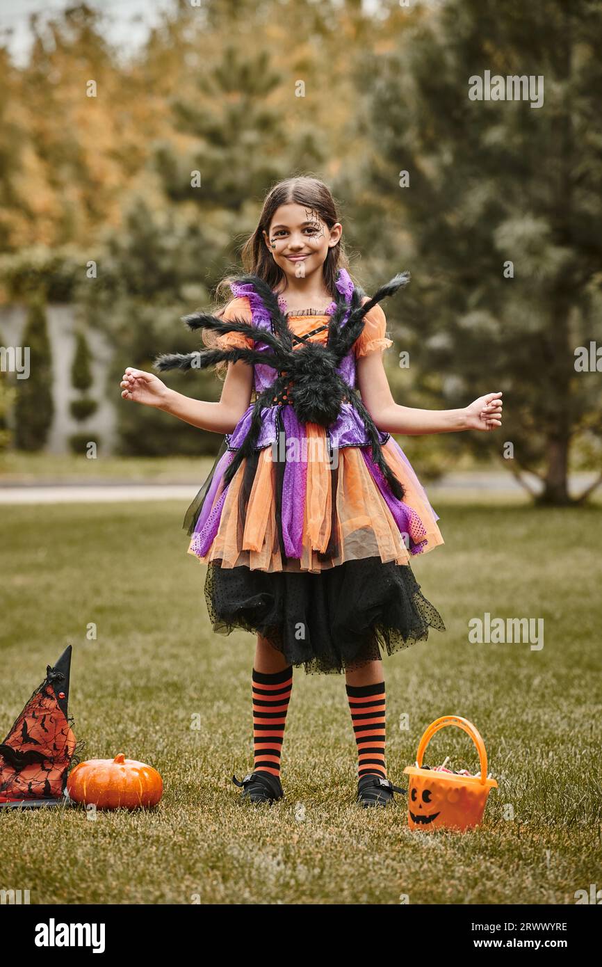 Glückliches Mädchen im Halloween Kostüm mit Spinne in der Nähe von Kürbis, Spitzhut und Süßigkeiteneimer Stockfoto