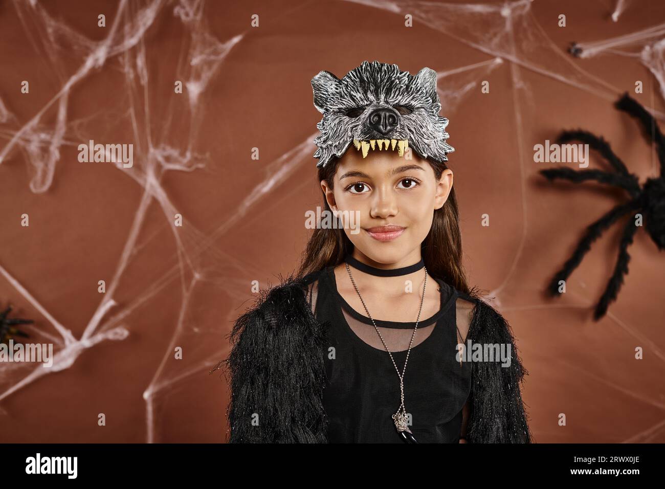 Niedliches Mädchen in schwarzem Outfit mit Wolfsmaske auf braunem Hintergrund, Nahaufnahme, Halloween-Konzept Stockfoto