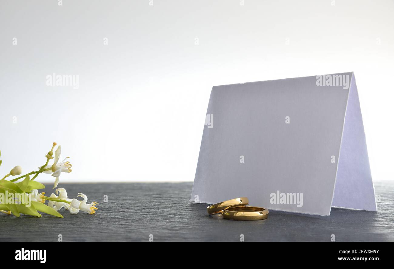 Zwei Verlobungsringe auf Schiefertisch mit leerer Papierkarte dahinter und Blumenstrauß und weißem isoliertem Hintergrund. Vorderansicht. Stockfoto