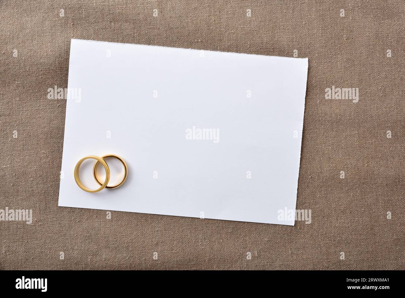 Weißer Hochzeitskartenhintergrund mit goldenen Ringen auf braunem Tuch. Draufsicht. Stockfoto